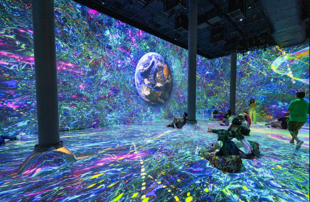 People sitting and standing inside a large room where the floor and walls are entirely covered by a projected image of Earth surrounded by dense, brightly coloured textures.
